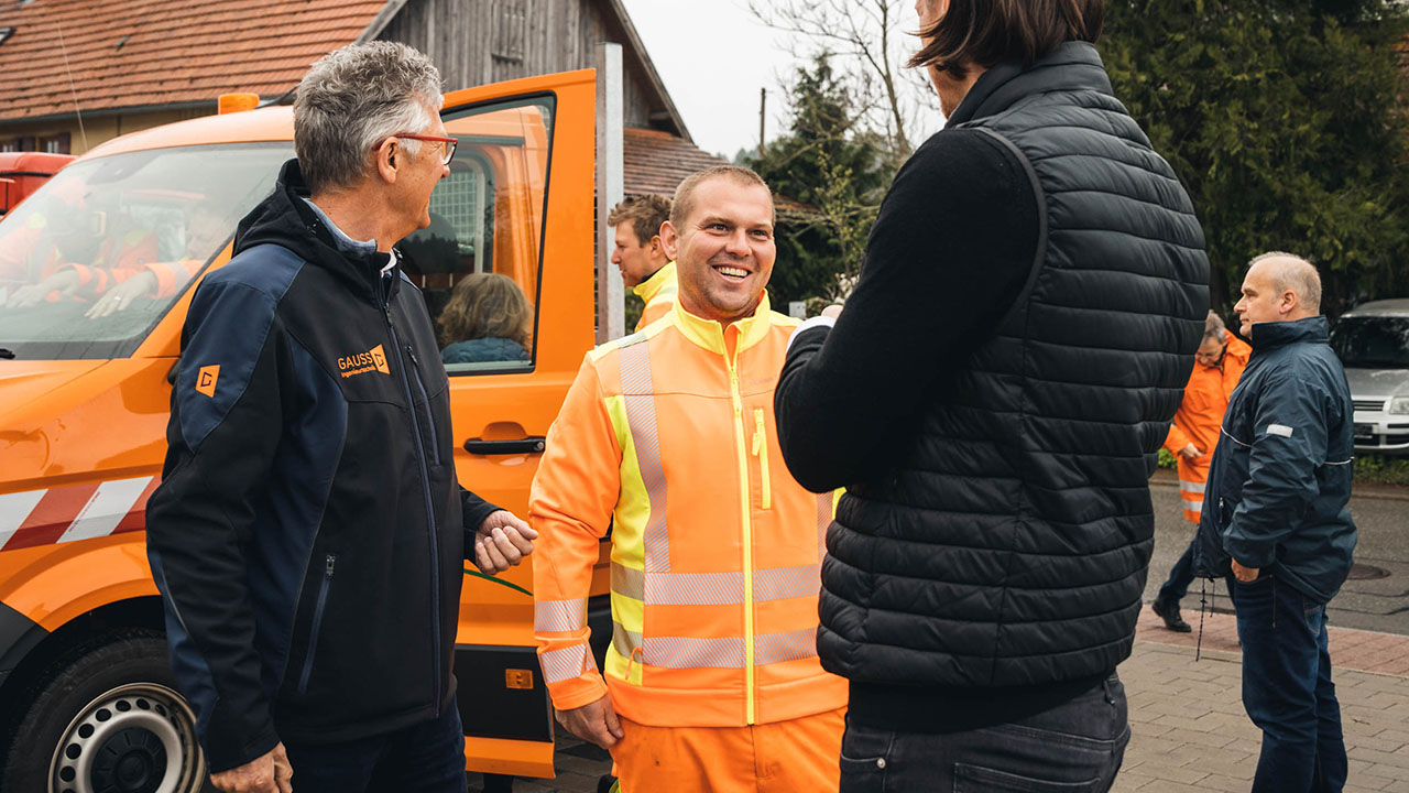 two men talking with a building yard worker in front of an orange car