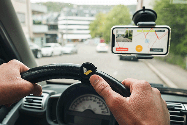 Two hands on a steering wheel while driving on a street. a bluethooth button on the steering wheel and a smartphone in a holder on the windshield with the vialytics street management system opened