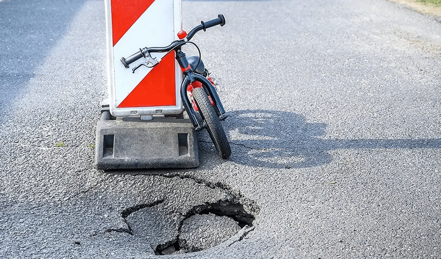 A child's bicycle leaning against a striped traffic barrier next to a deep pothole in the asphalt, illustrating the dangers of poor road surface conditions that lead to bicycle accidents.